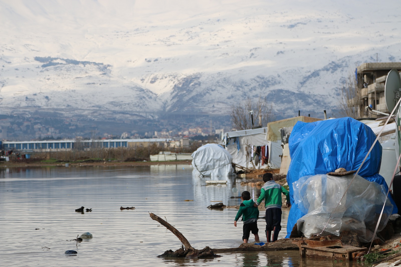 Children walk through a landscape of water, makeshift tents, and huge snow-covered mountains dominating the background.