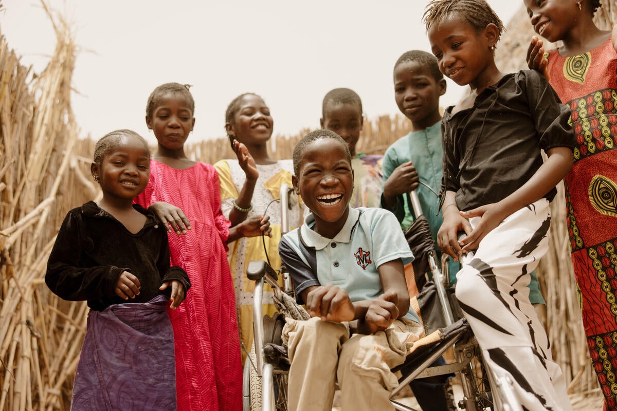 A boy in a wheelchair beams with a joyful smile while surrounded by children. Tall, dry stalks line the background.