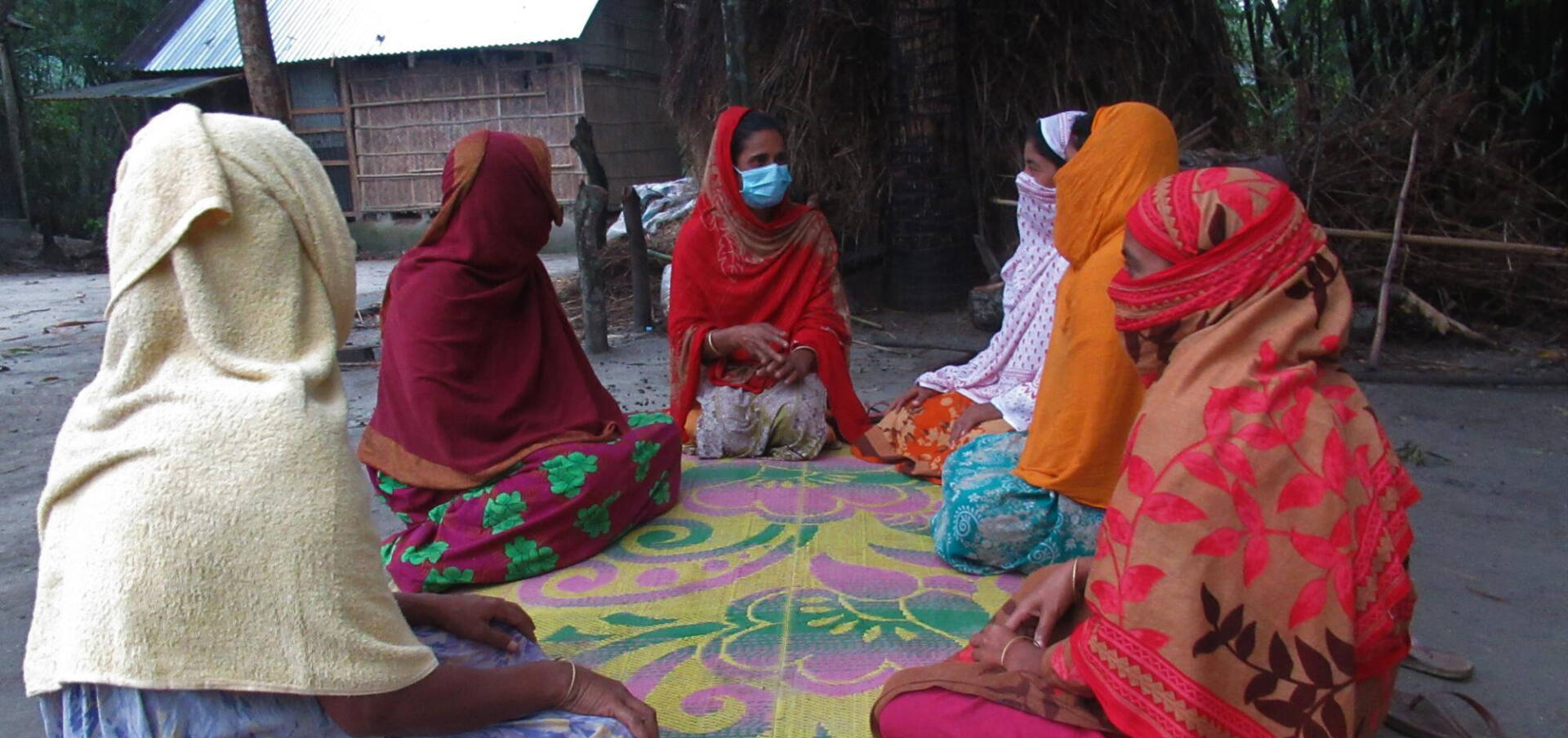 Six Bangladeshi women in colorful attire sit on a colored mat outside.
