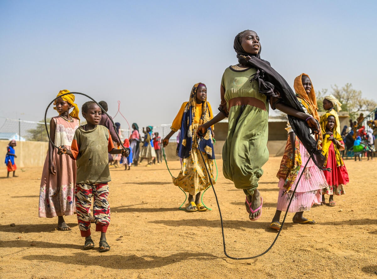 A girl in a headscarf and green dress jumps rope, mid-air, surrounded by other children jumping rope.