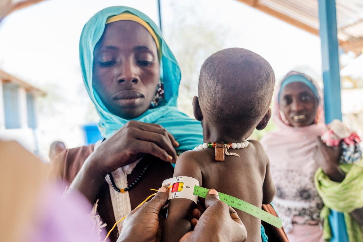 A woman in a turquoise headscarf holds her young child while a health worker measures the child&rsquo;s arm for malnutrition. Another woman looks on in the background.