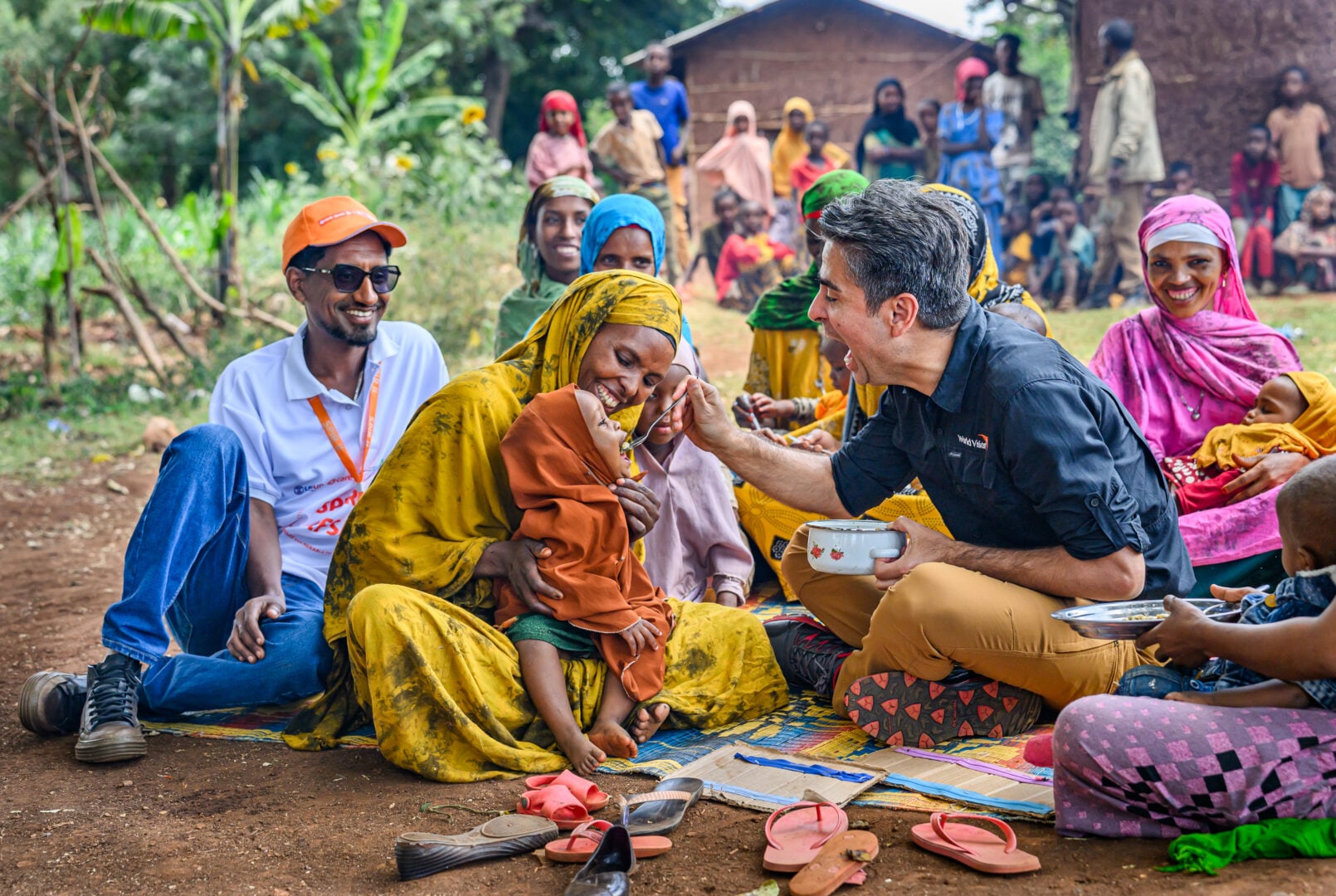 A man feeds a young girl sitting on her mother’s lap. She mimics his open mouth as a spoon comes her way, while surrounded by laughing community members outdoors.