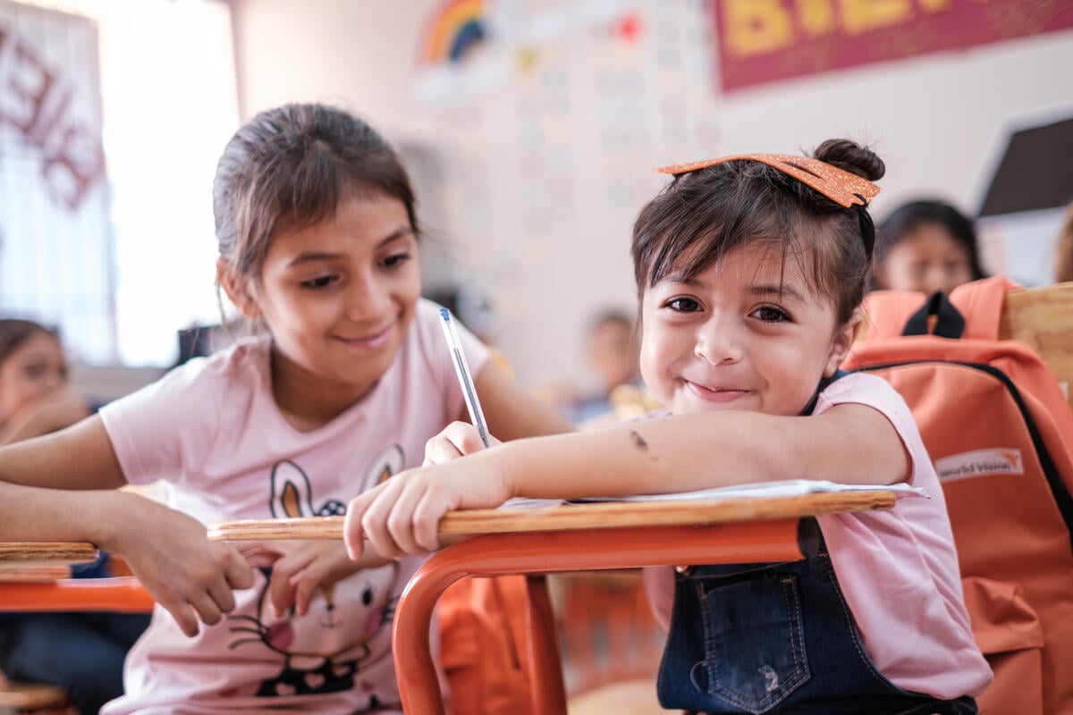 Two young girls in pink T-shirts smile brightly in a classroom setting. One girl looks at the camera, while the other gazes at her classmate.