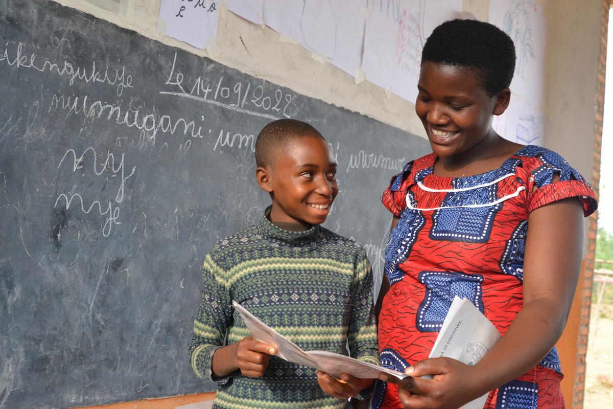 A girl and woman smile at one another while together holding open a booklet. A blackboard with writing on it is behind them.