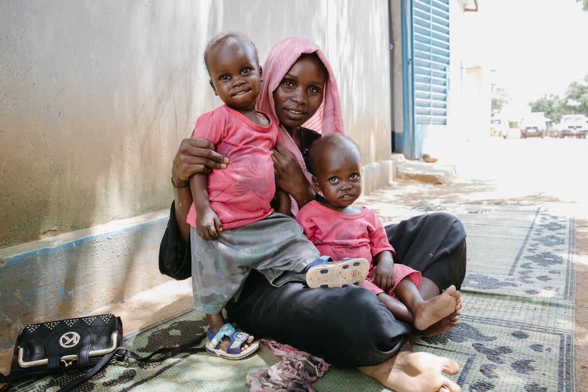 A woman in a pink headscarf sits on the ground on a blue carpet, gazing into the camera as she holds a young boy in her arms. The boy&rsquo;s twin, wearing a matching pink shirt, sits in her lap. 
