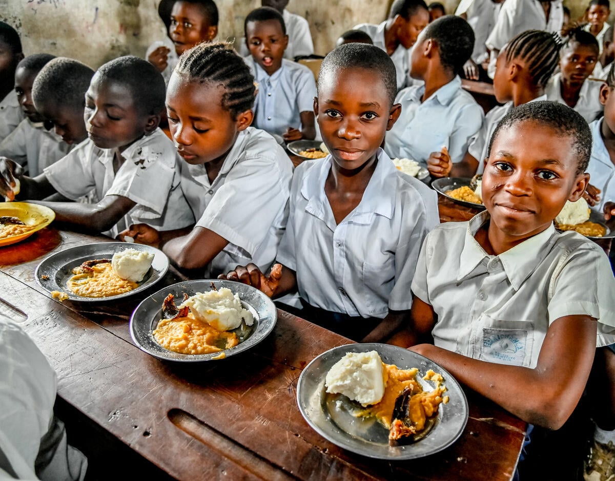 Girls in school uniforms sit in rows in a classroom, each with a plate full of food.