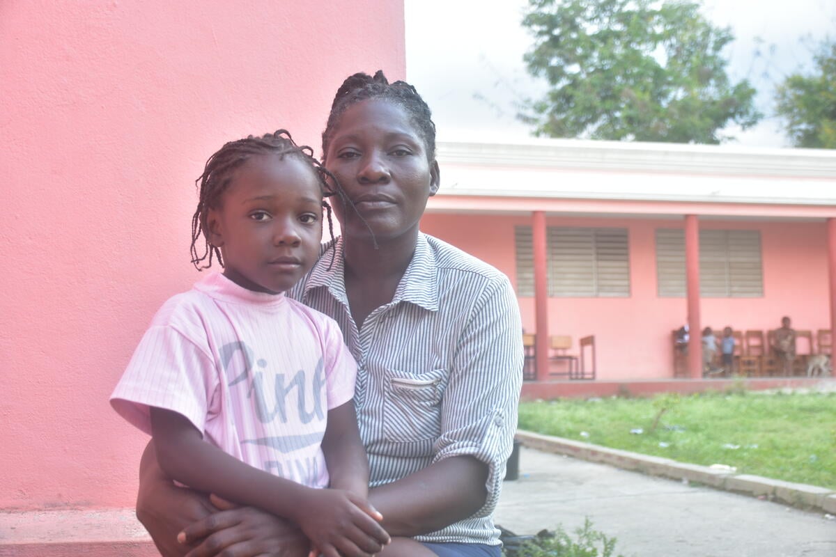 A young girl in a pink shirt sits on a woman&rsquo;s lap, both gazing at the camera in front of a pink building.