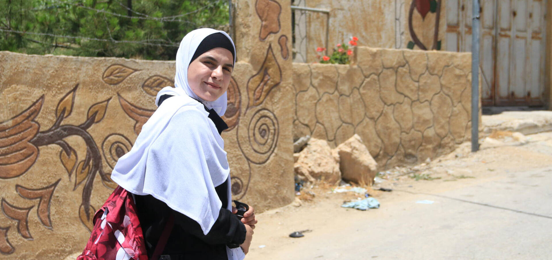 A girl in a white headscarf walks forward while looking back at the camera.