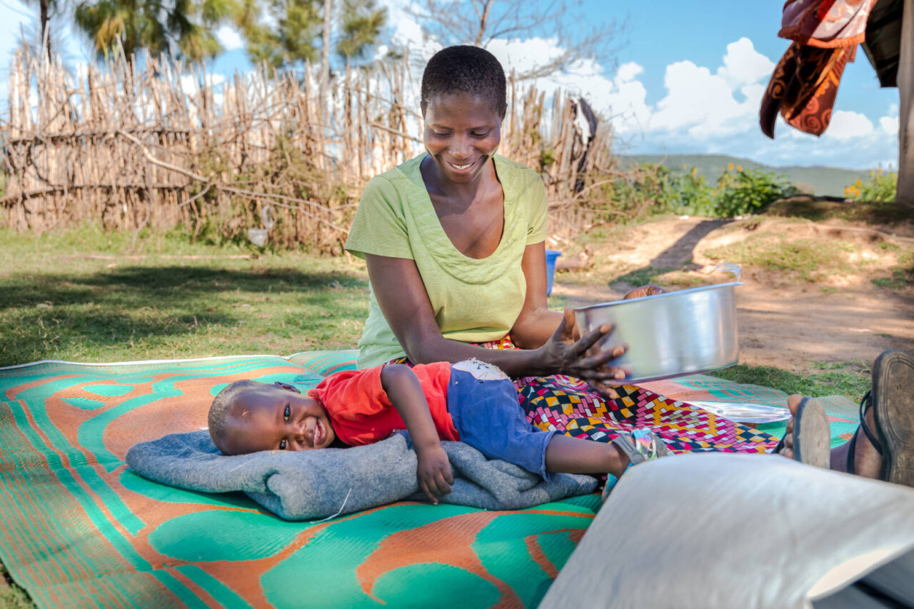 Sitting on a colorful blanket, a mother holds a silver bowl while looking down on her young son hugging a blanket.
