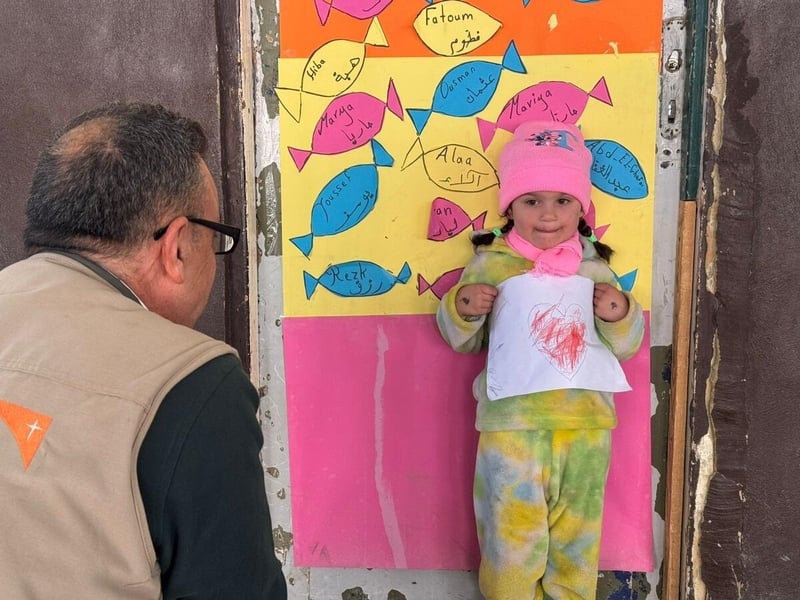 A young girl holds up a drawing of a heart while standing against a colorful backdrop. A man wearing a World Vision vest looks at her while kneeling down to her eye level.