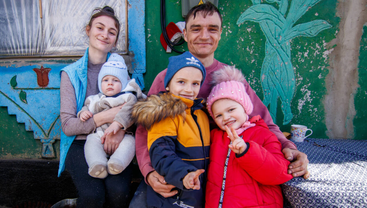 A family in Moldova sits. Dressed in winter gear, the father embraces his two children while the mother holds a baby.