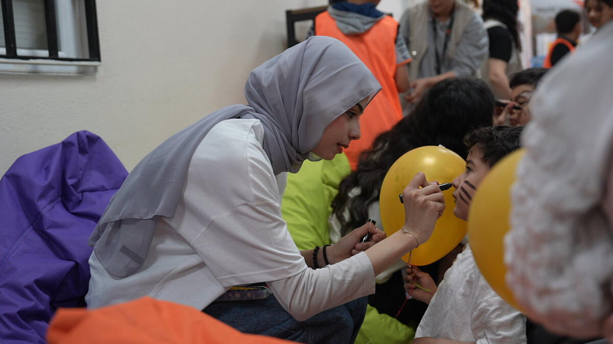 A girl leans forward from her chair to draw on a young boy&rsquo;s face, surrounded by balloons and people.