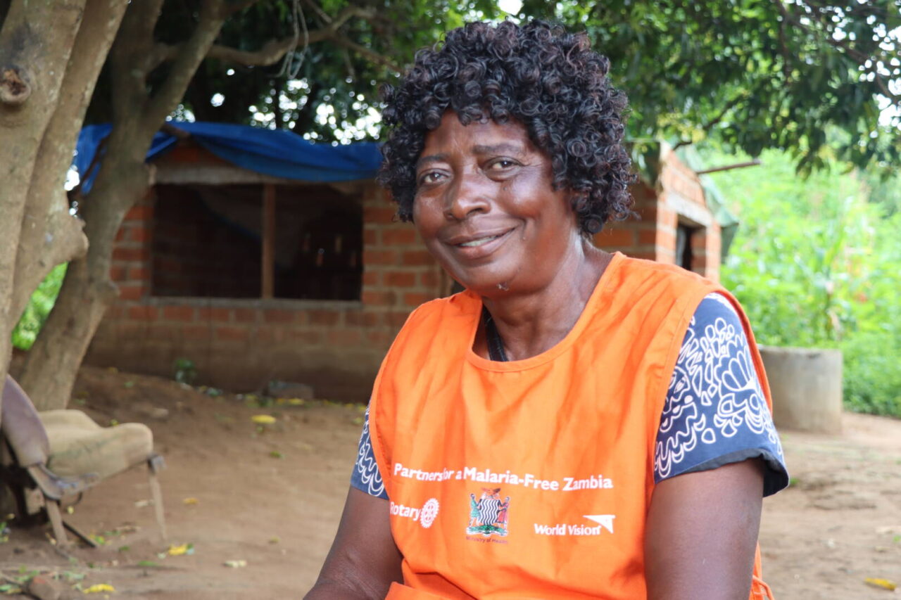 A woman is seated under a tree, wearing an orange World Vision vest that reads &ldquo;Partners for a Malaria-Free Zambia.&rdquo;