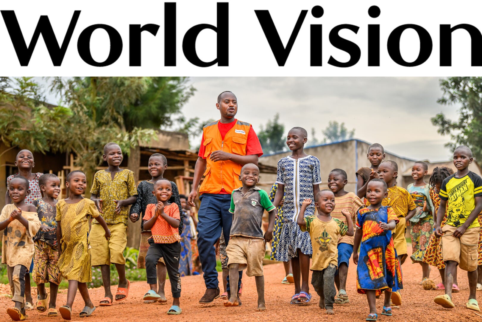 A group of smiling children walk with a World Vision staff member wearing an orange vest on a dirt road.